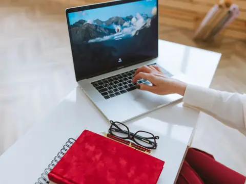 A laptop, a red notebook and glasses on a bright white desk