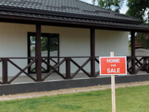 A white single-story home with a wide porch and a red Home for Sale sign on the lawn