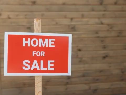 A red Home for Sale sign on a wooden post against a wood wall