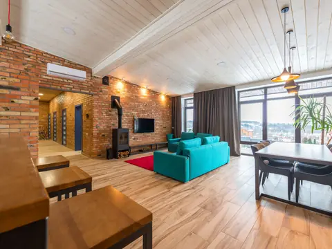 A sunny living room with a brick wall, a teal sofa, a wood stove and a dining table by tall windows