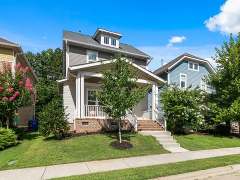 A two-story home with a front porch, a young tree and flowering shrubs on a sunny day