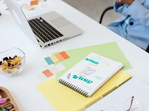 An open sketch notebook beside a laptop and color cards on a white desk